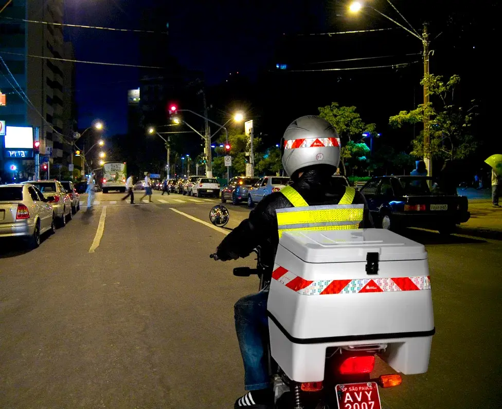 Motociclista com faixa refletiva no baú de moto. O motociclista pilota durante a noite, em uma cidade.